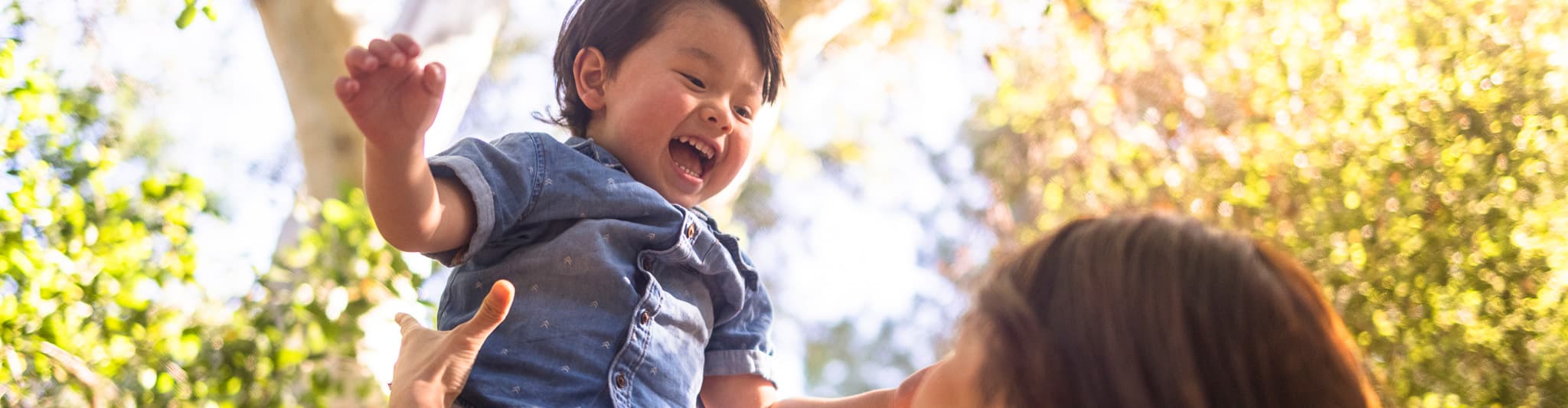 Laughing toddler being lifted up in the air by his mother amongst the trees